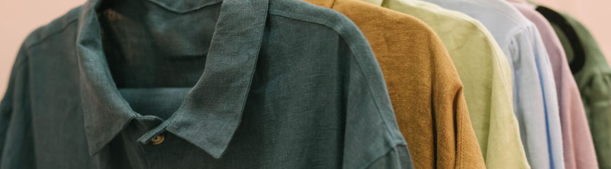 Rack of collared button-front shirts in teal, ochre, sage and pale blue, with the teal linen shirt nearest showing its collar, placket and buttons